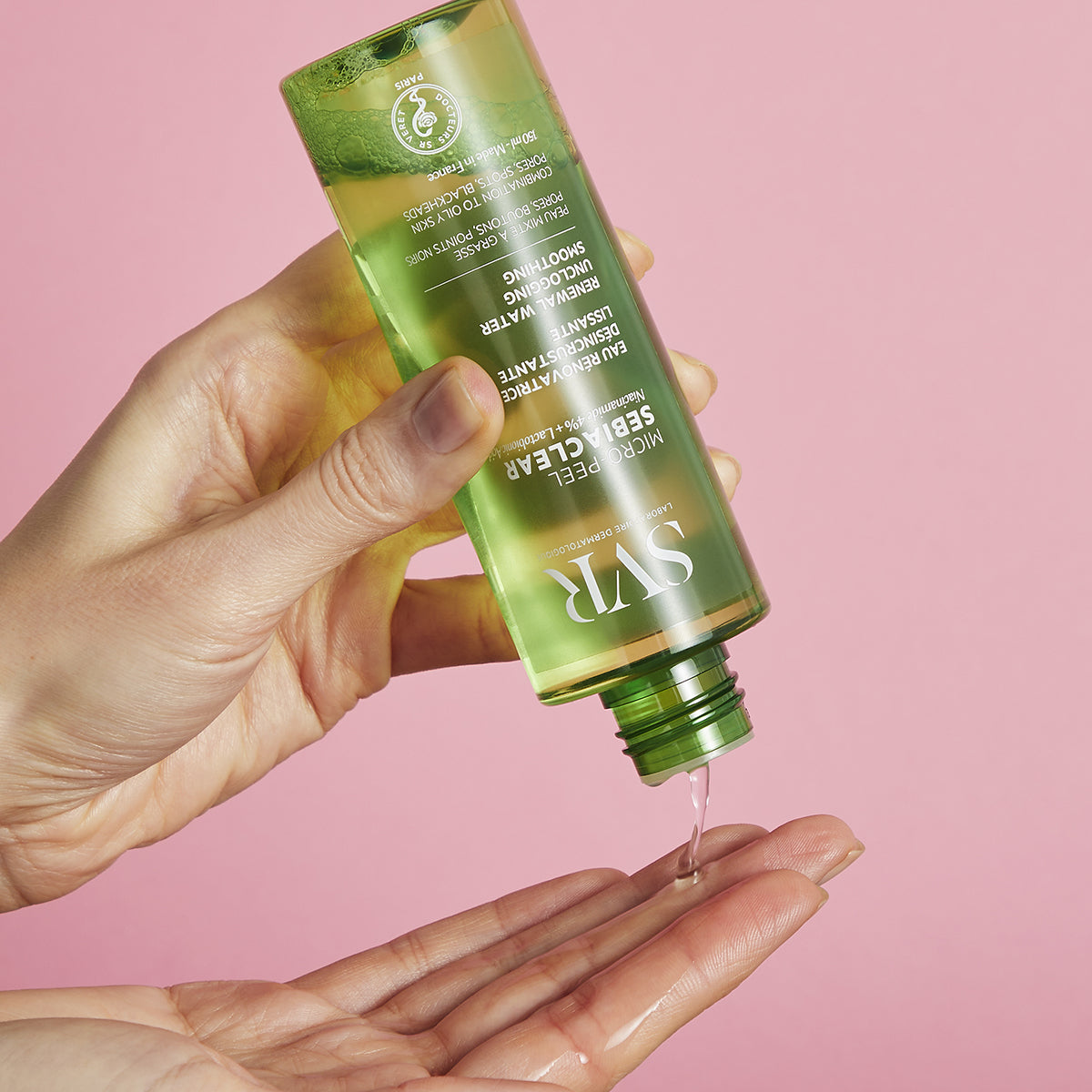 Clear green bottle of SVR Sebiaclear Micro-Peel pouring liquid onto a hand against a soft pink background.