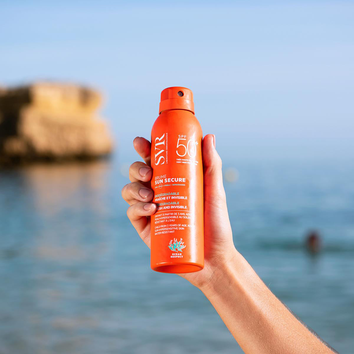 Hand holding an orange SVR SUN SECURE SPF50 sunscreen spray bottle against a blurred beach and ocean background.