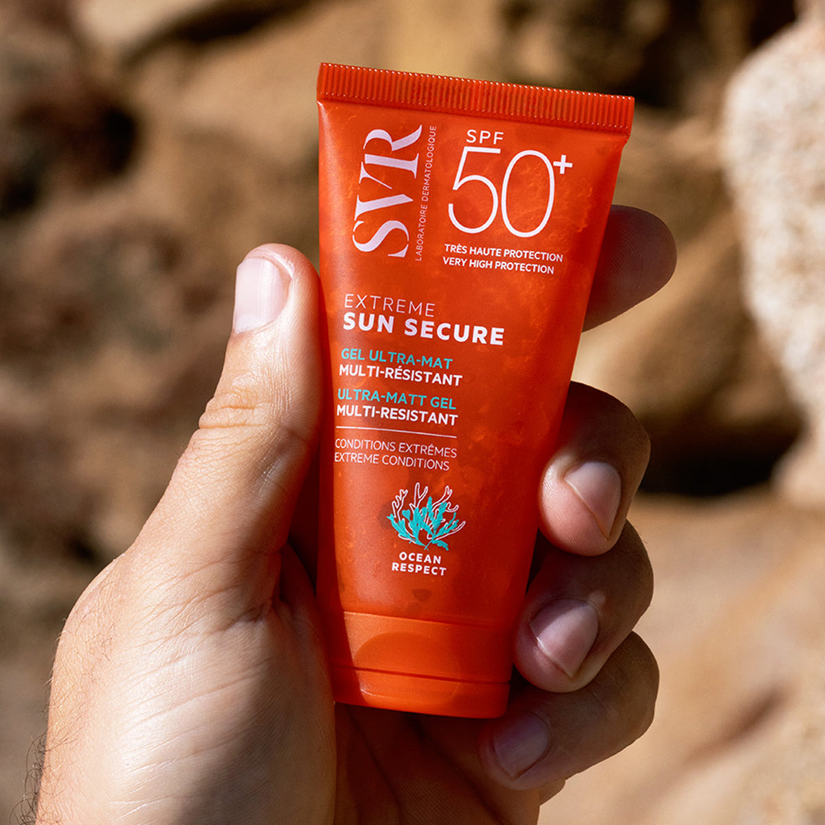 Hand holding an orange SVR SUN SECURE Extrême SPF50+ ultra-matt gel sunscreen tube against a rocky background.