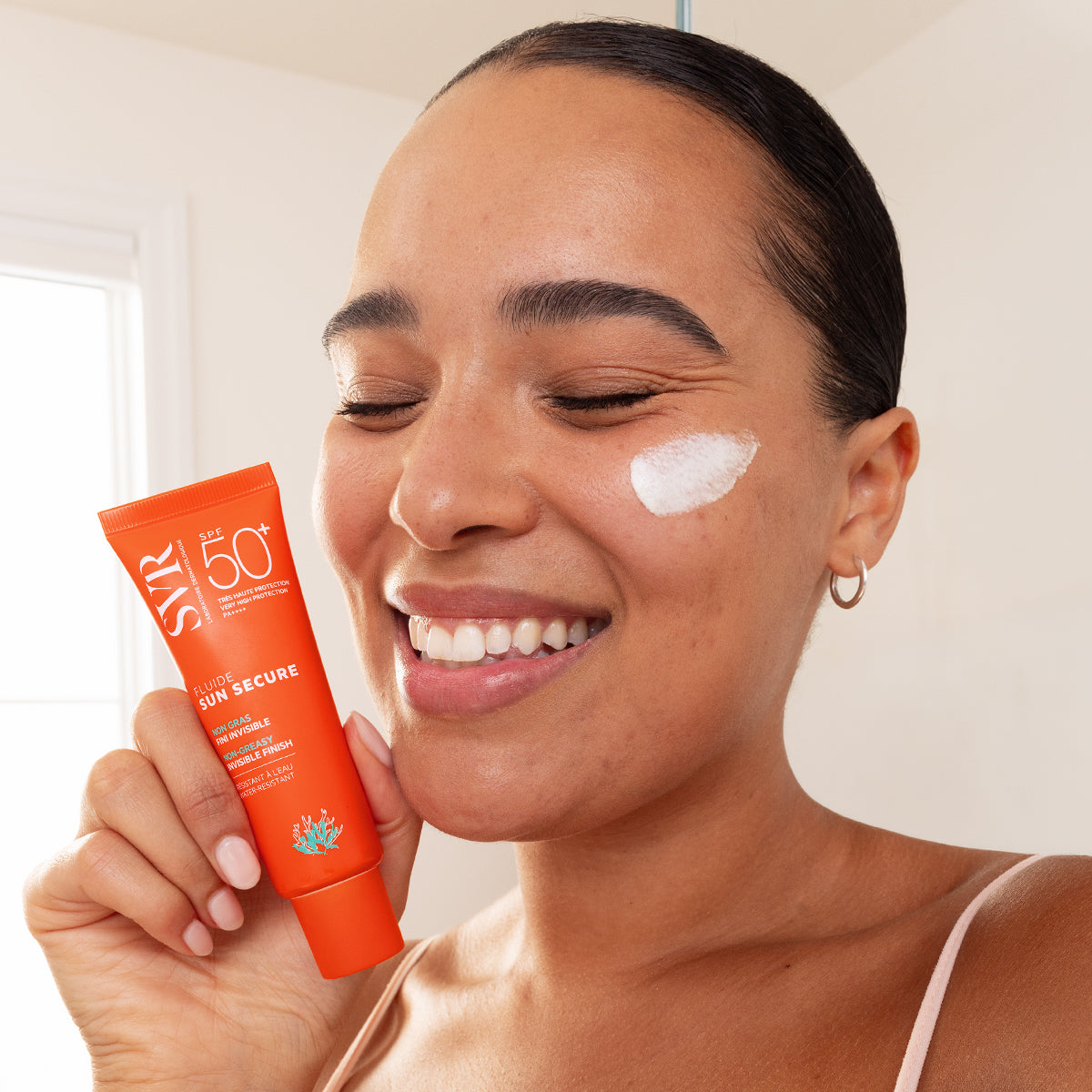 Woman holding an orange SVR SUN SECURE SPF50+ sunscreen tube with a dab of white cream on her cheek.