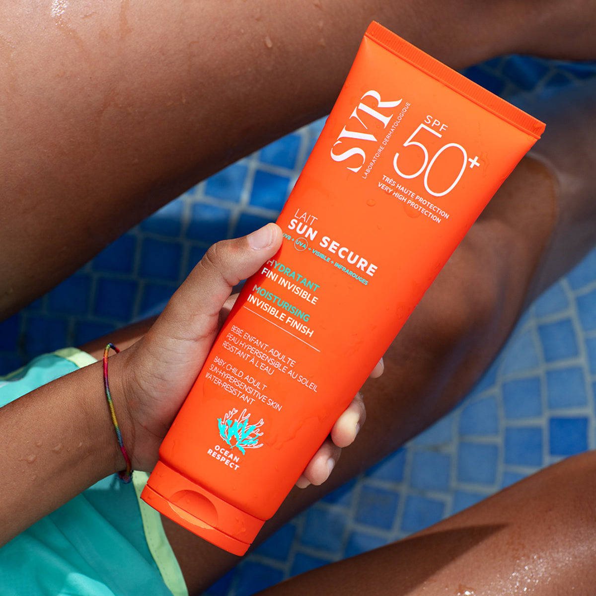 Bright orange SVR SUN SECURE SPF50+ sunscreen tube held by a person near a blue tiled pool.