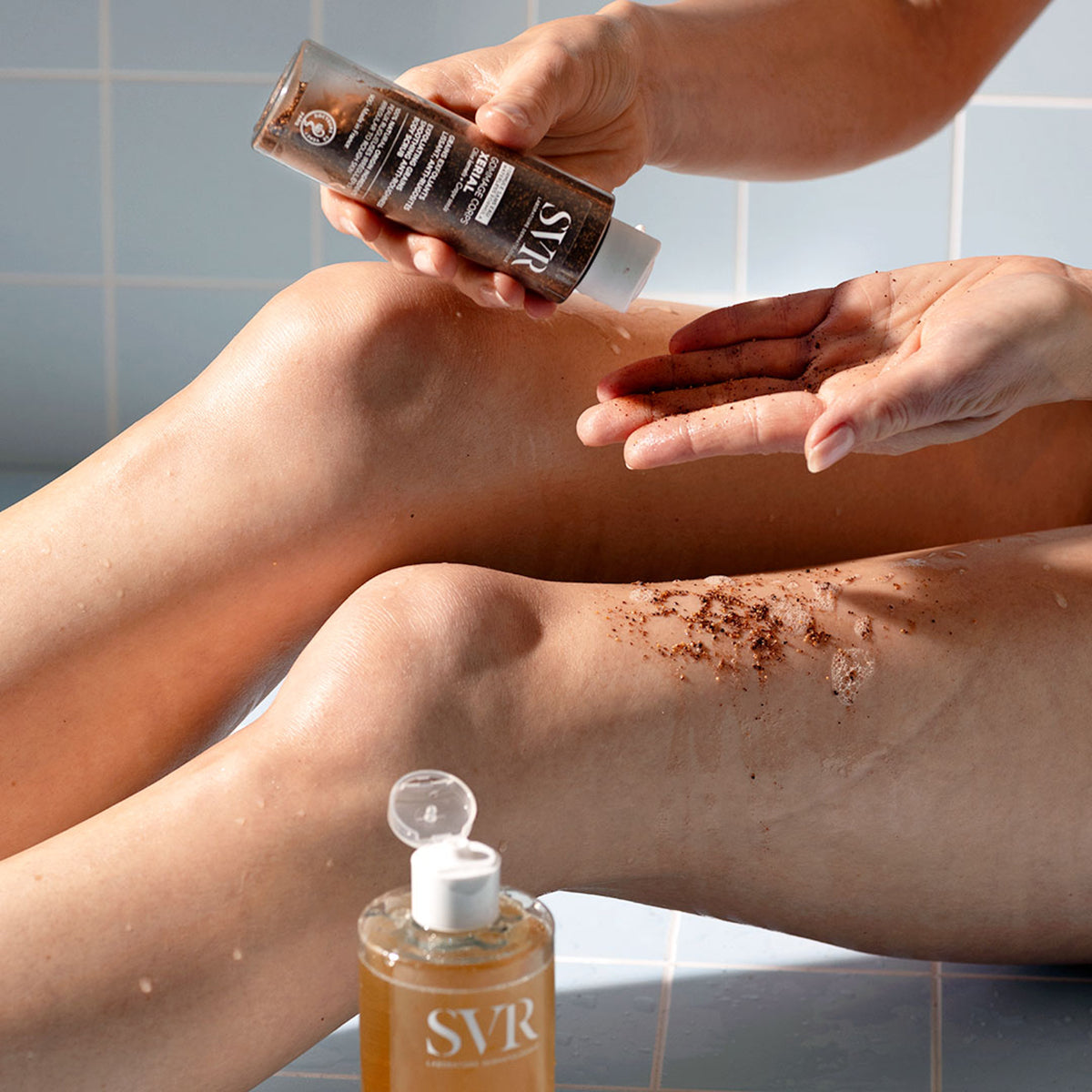 SVR XERIAL Gommage body scrub being applied to a leg, showing the brown exfoliating texture and clear plastic bottle.