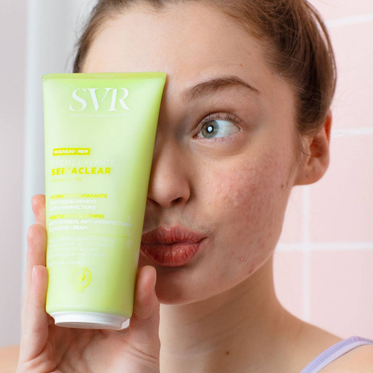 Woman holding a light green SVR Sebiaclear Creme Lavante tube against her face in a bathroom setting.
