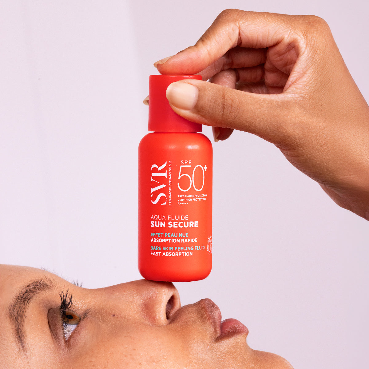 Hand holding a bright orange SVR SUN SECURE Aqua Fluide SPF50+ sunscreen bottle above a person's nose.