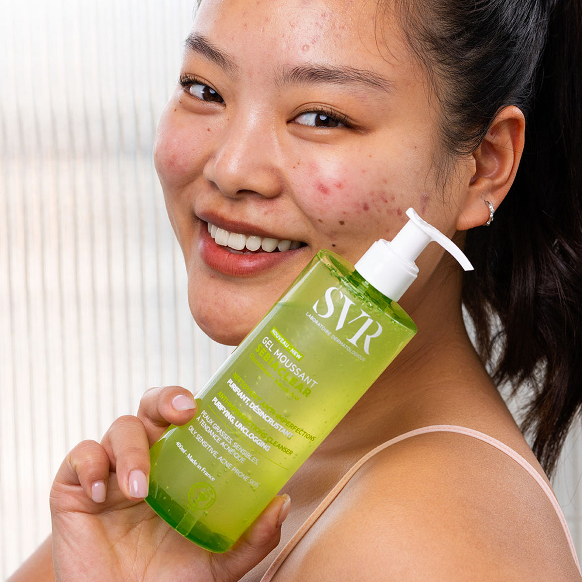 Smiling woman holding a 400ml bottle of green SVR Sebiaclear Gel Moussant cleanser with a white pump dispenser.