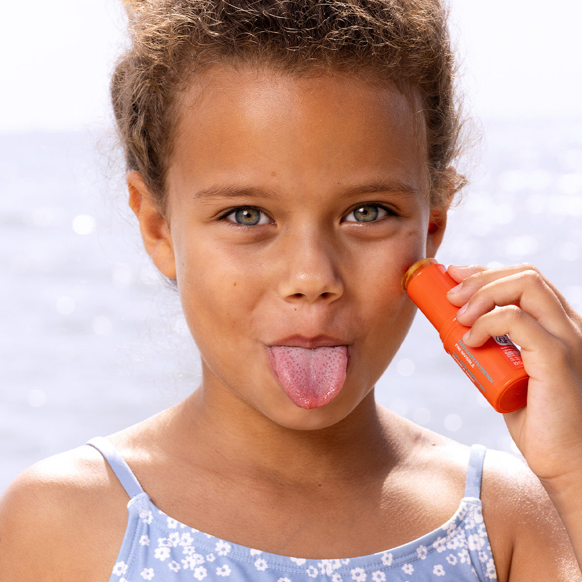 Young girl applying orange SUN SECURE SPF50+ sunscreen stick to her cheek against a bright, sunny ocean background.