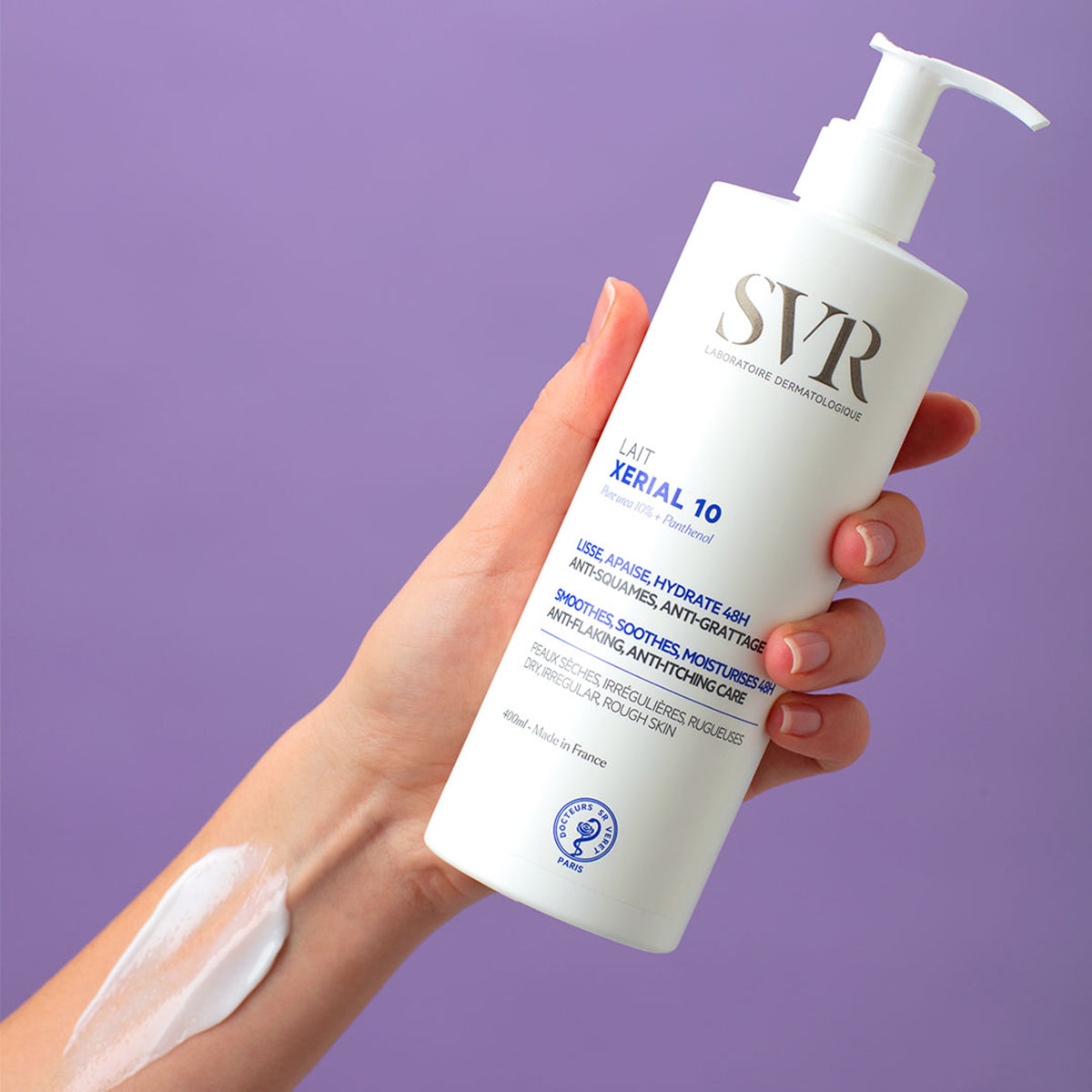 Hand holding a 400ml white pump bottle of SVR XERIAL 10 Lait body lotion against a purple background with cream on the arm.