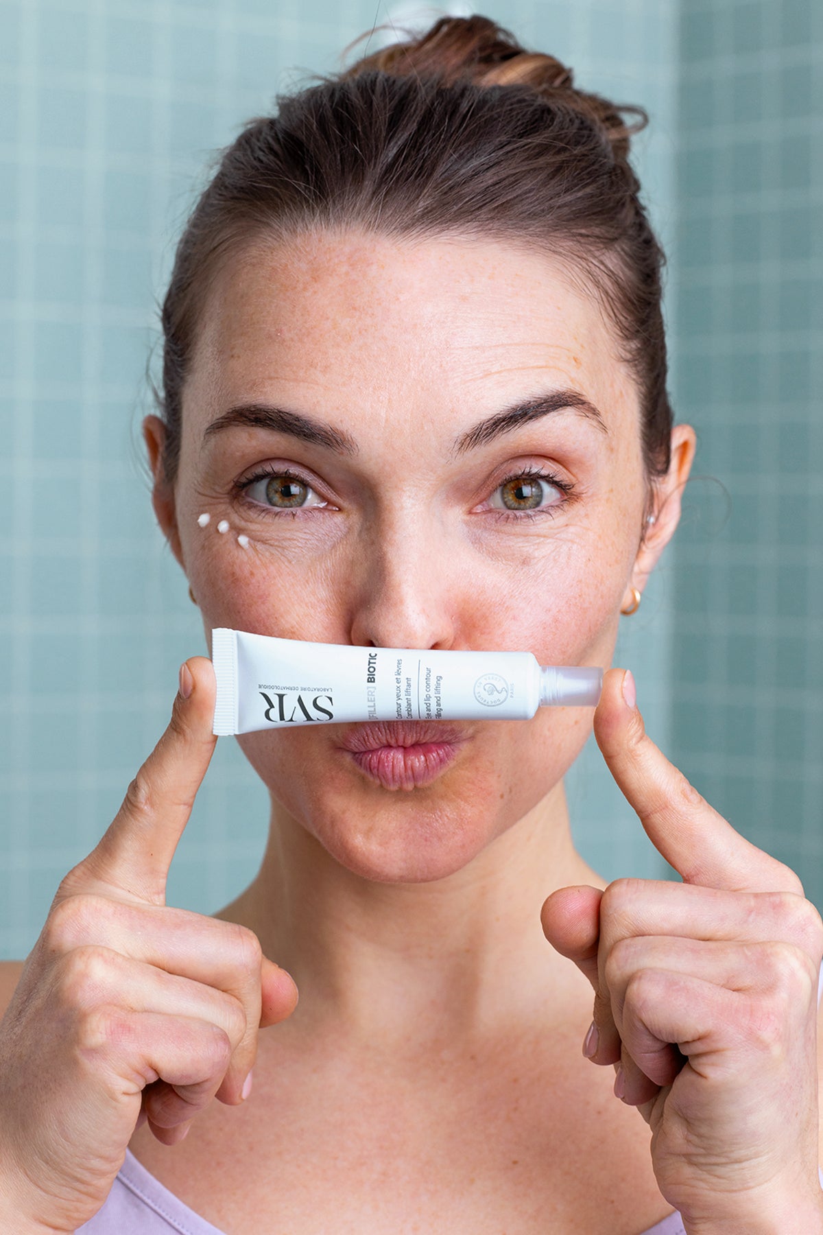 Woman holding a white SVR Filler Biotic eye and lip contour cream tube in front of her lips with cream dots on her face.