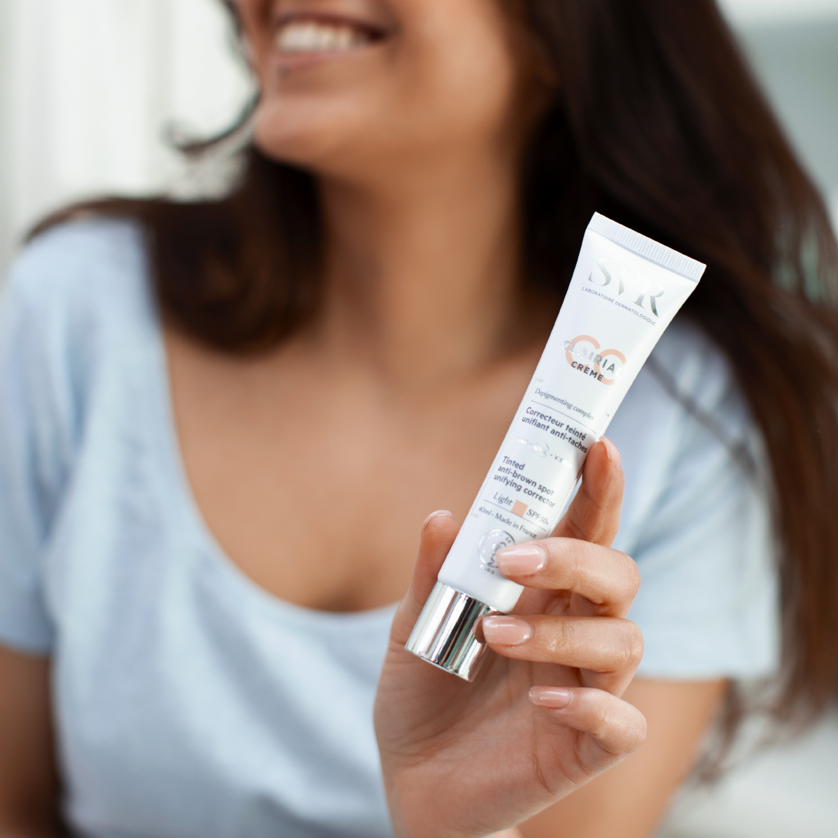 Woman holding a white 40ml SVR Clairial CC Light Crème SPF50+ tube with a silver cap against a blurred background.