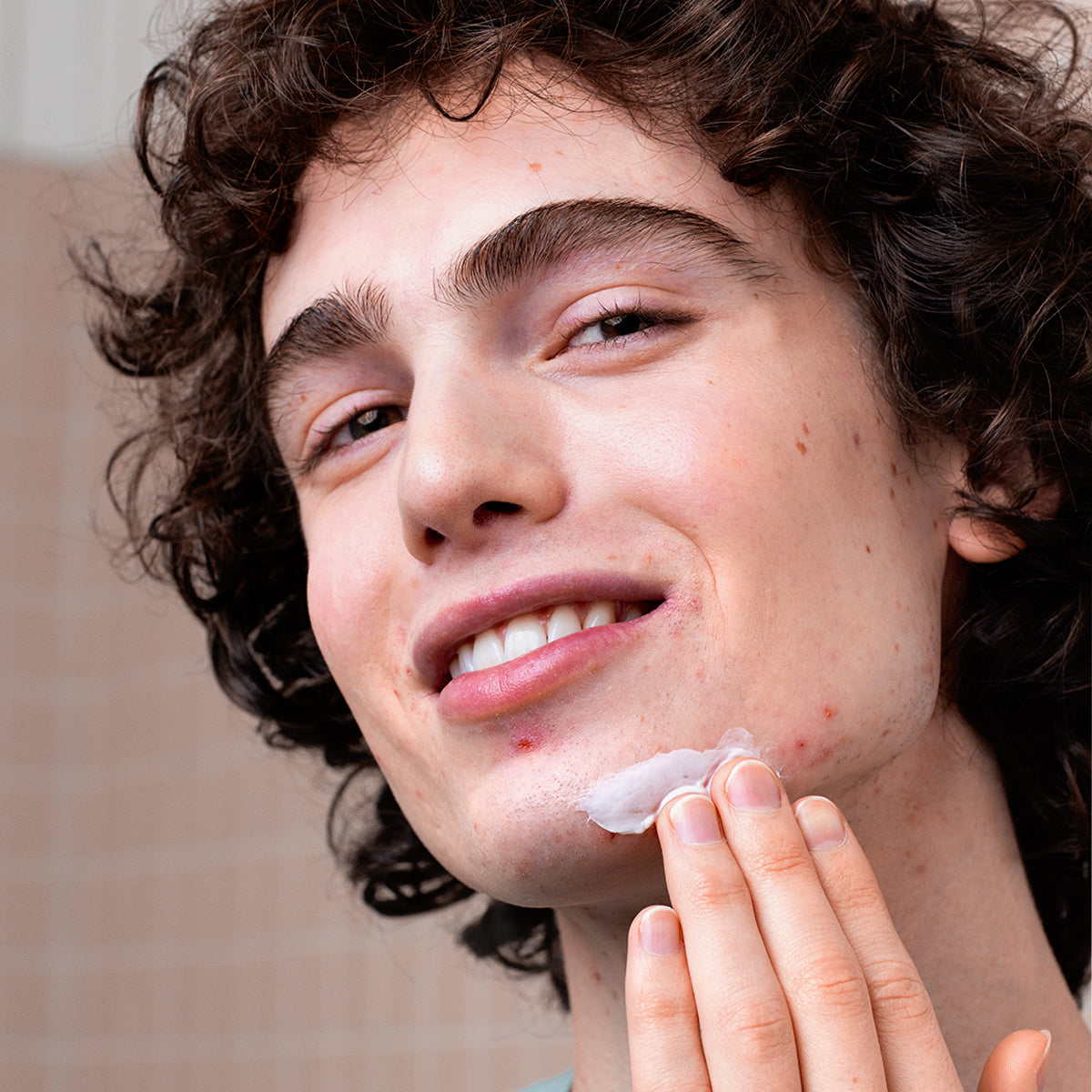 Young person with curly hair applying white CICAVIT+ Crème HPPI to a blemish on their chin in a bathroom setting.