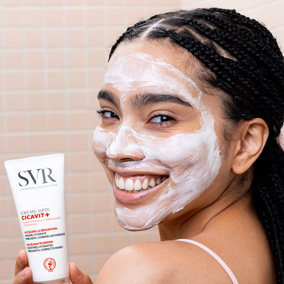 Smiling woman with white CICAVIT+ Crème HPPI applied to her face, holding the white skincare tube against a tiled background.