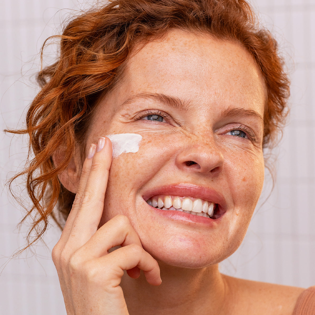 Smiling woman with red hair applying white SUN SECURE SPF50+ fluid cream to her cheek against a tiled background.