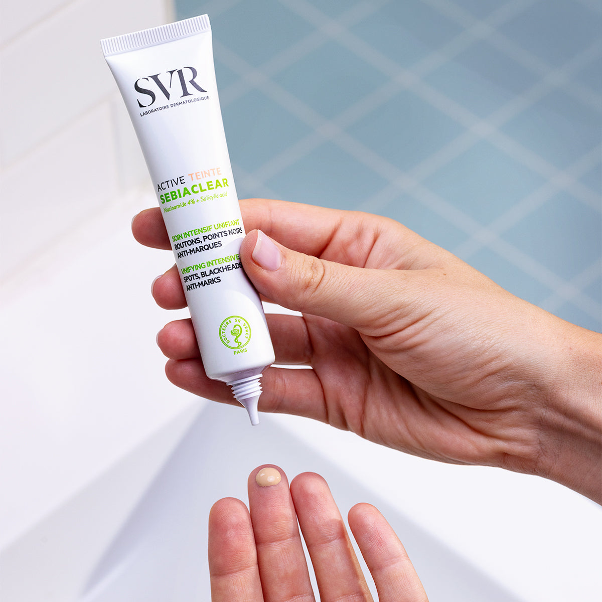 Hand holding a white SVR Sebiaclear Active Teinte tube, dispensing a drop of tinted facial cream onto a fingertip.