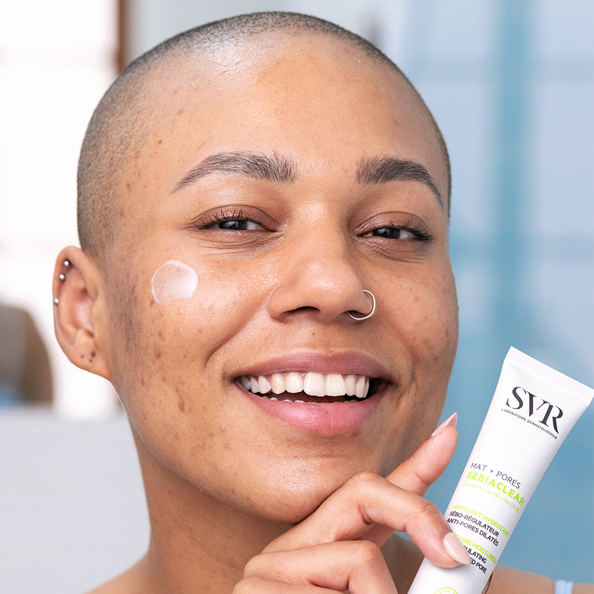 Smiling person with a dollop of SVR Sebiaclear Mat+Pores cream on their cheek, holding the white tube of skincare.